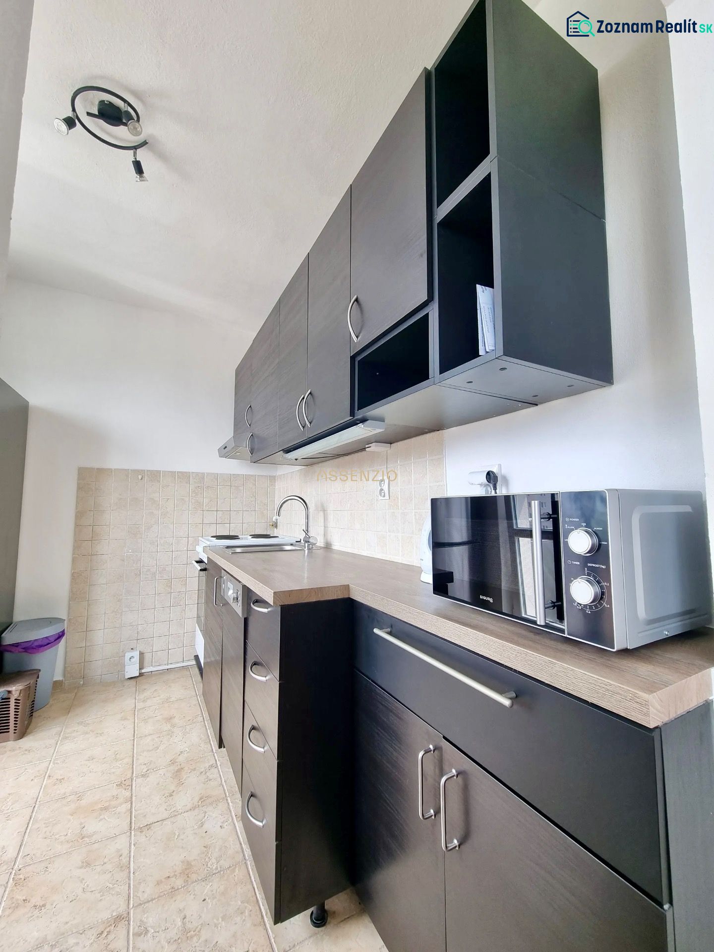 A kitchen in a 2-room apartment with dark cabinets and a microwave, floor with wood decor.