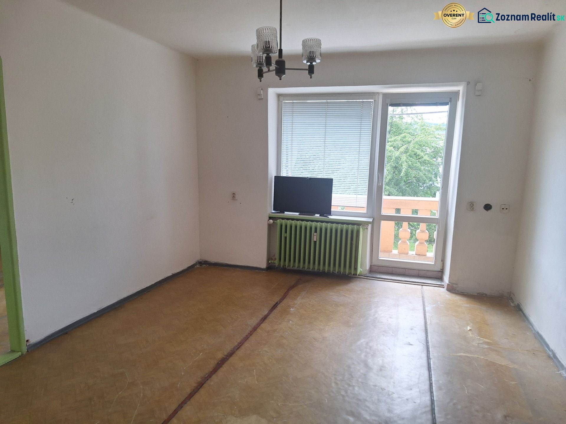 An empty room in a two-room apartment with a wood-patterned floor and a window to the balcony.