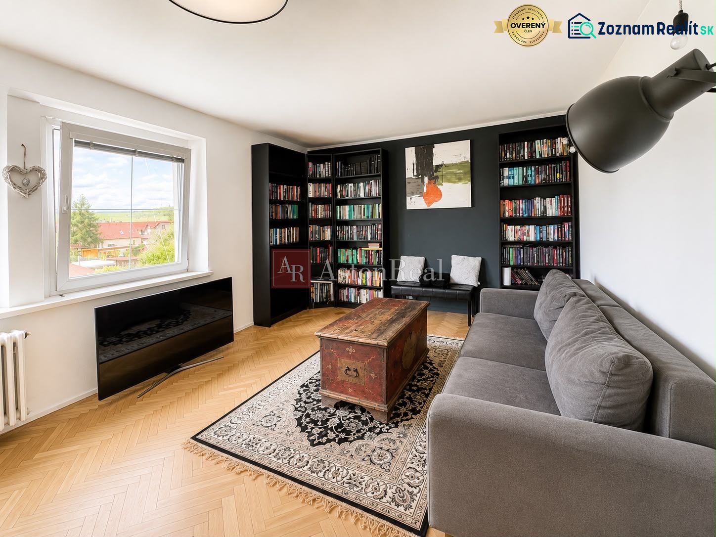 Living room in a 3-room apartment with a sofa, bookshelf, wood-patterned floor, and a large window.