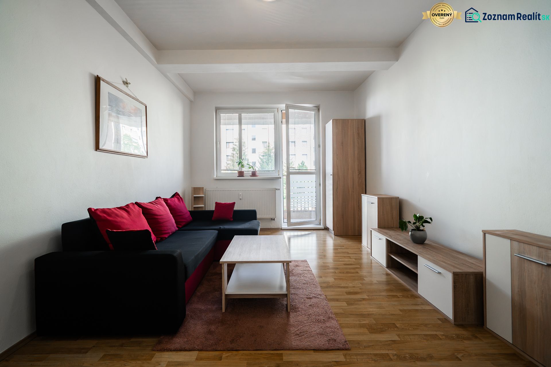 Living room in a two-room apartment with a sofa, red cushions, and a wooden-patterned floor.