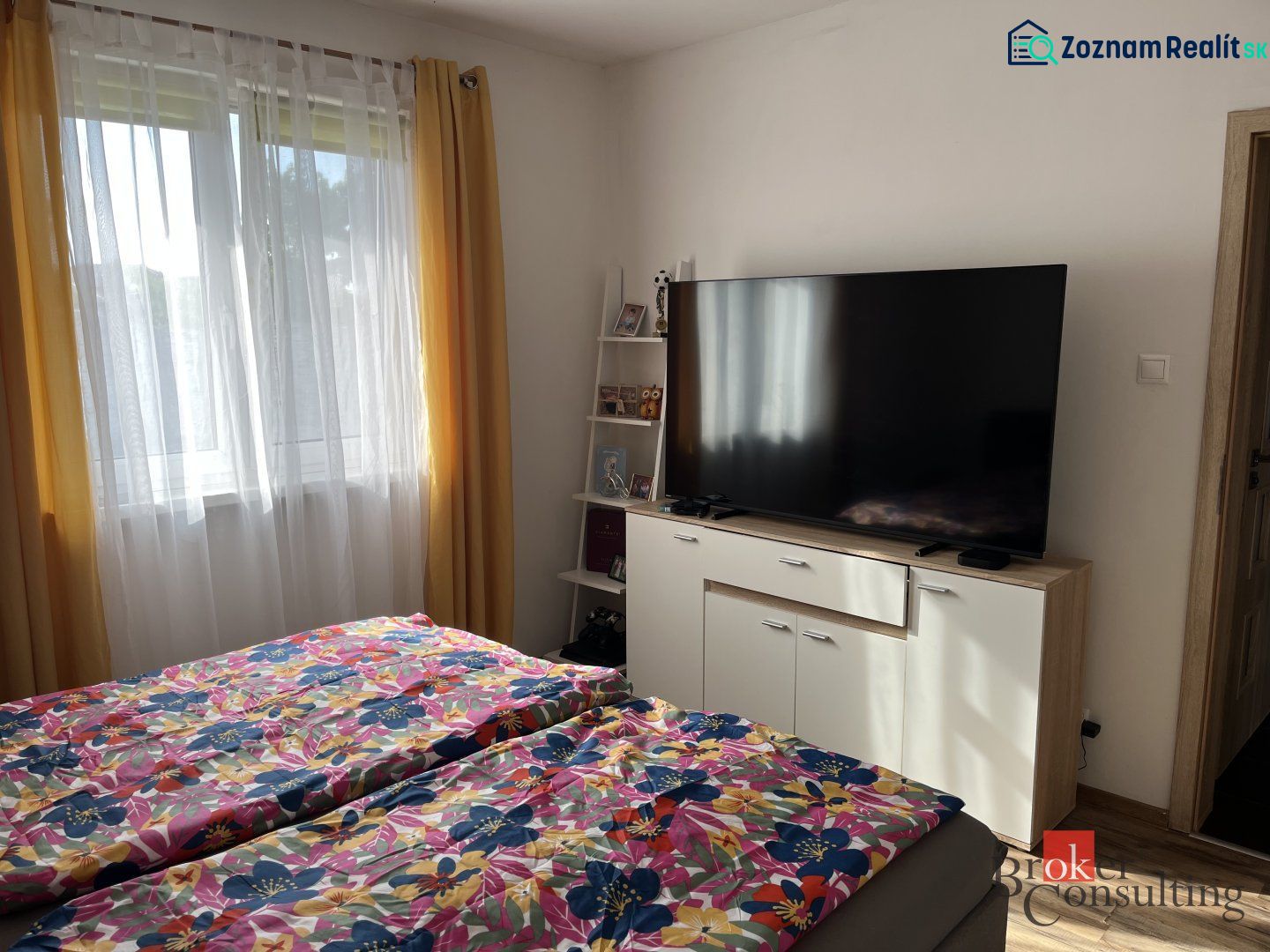Bedroom in a family house with TV, floral bed linens, yellow curtains, window.