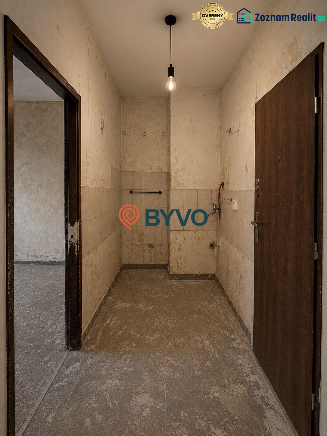 A spacious entrance area in a studio apartment with older tiles and lighting by a bulb.