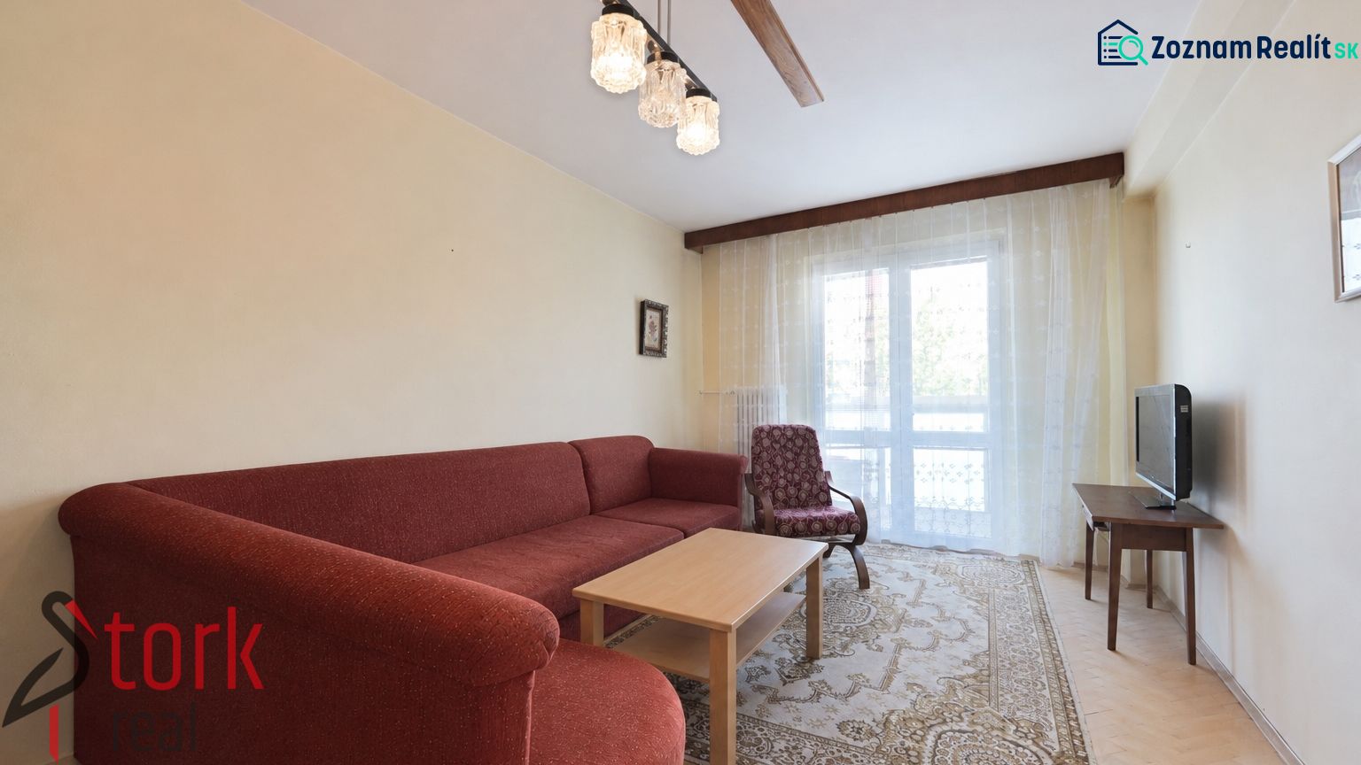 Living room of a three-room apartment with a red sofa, TV, carpet, and wooden decor flooring.