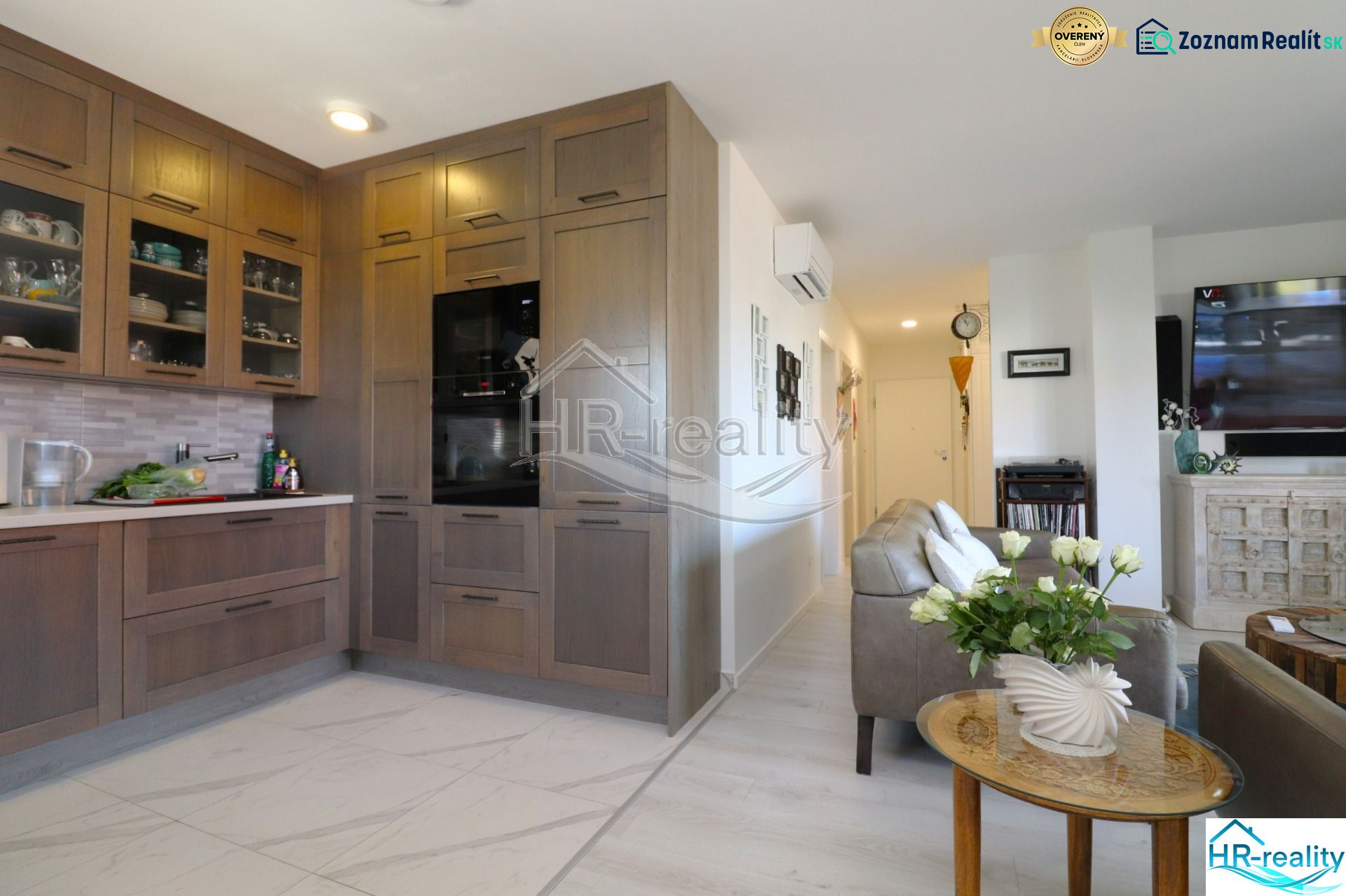 A kitchen in a holiday apartment with an open living room, wooden cabinets, and light flooring.