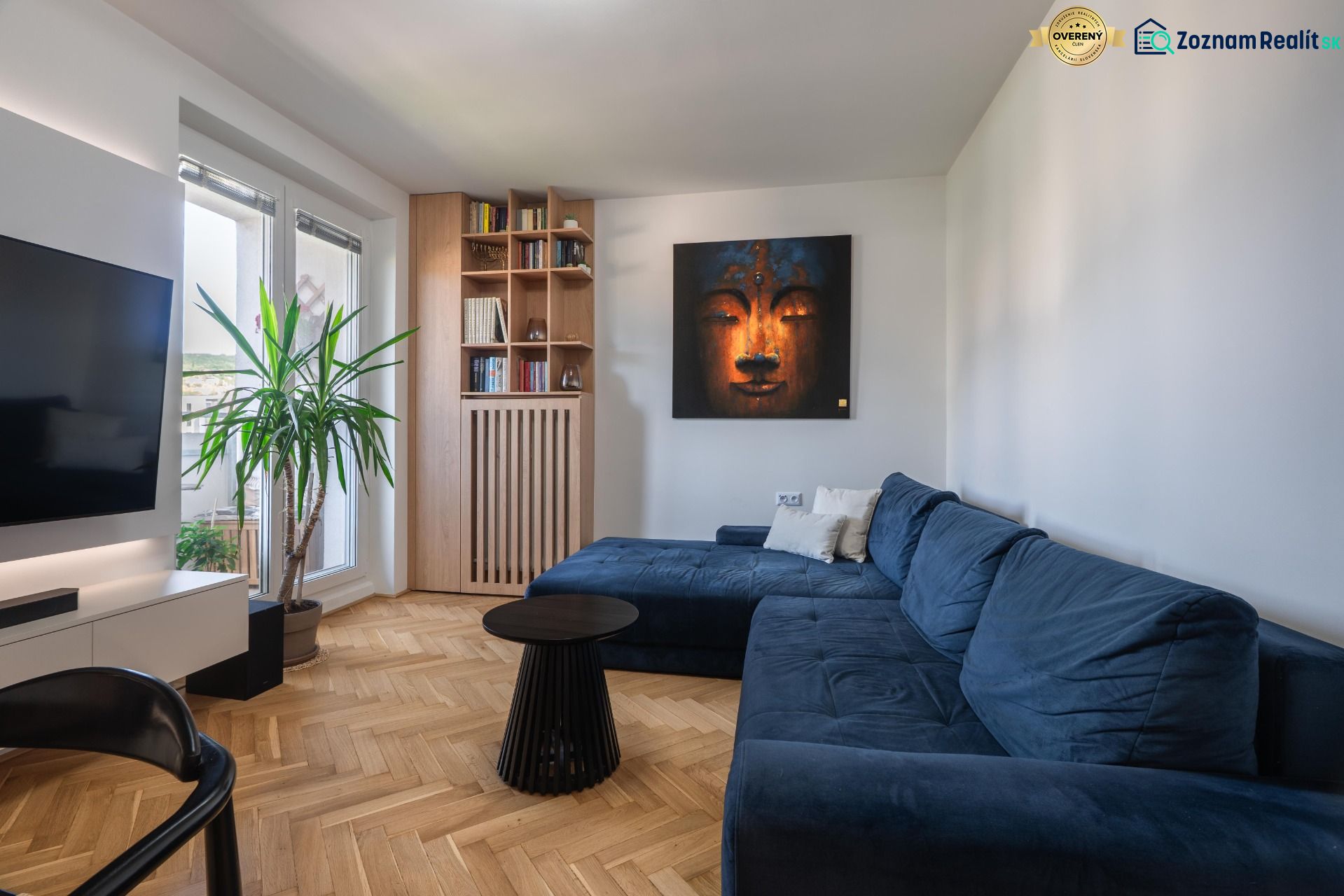 Living room with a blue sofa, painting, bookshelf, and wooden decor flooring in a three-room apartment.