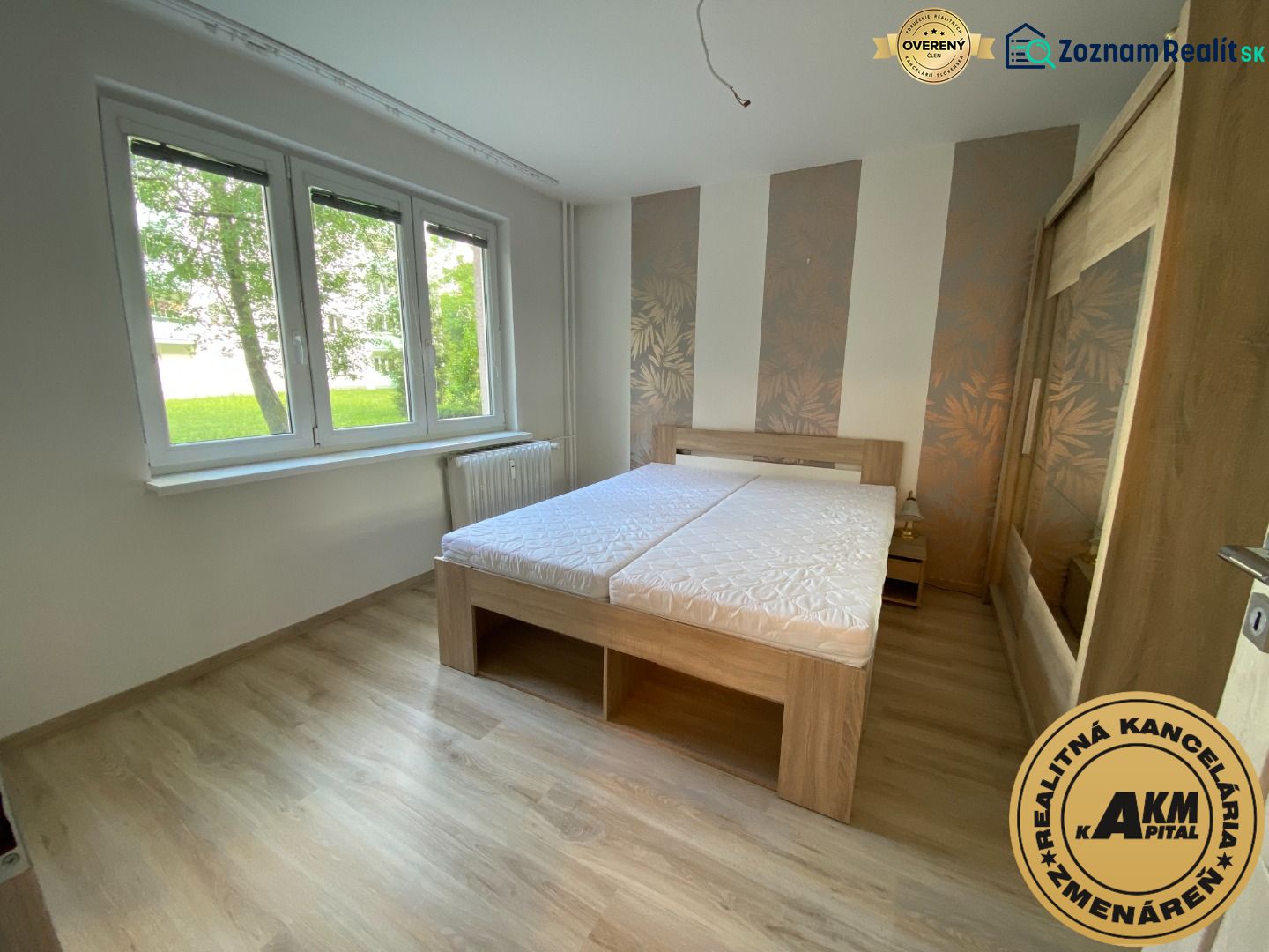 A bedroom in a two-room apartment with a wood-patterned floor and large windows.