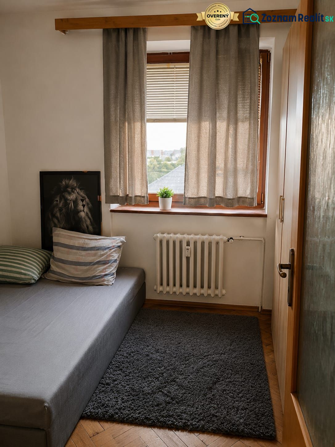 A room in a 3-room apartment with a bed, curtains, and a carpet on a floor with a wooden decor.
