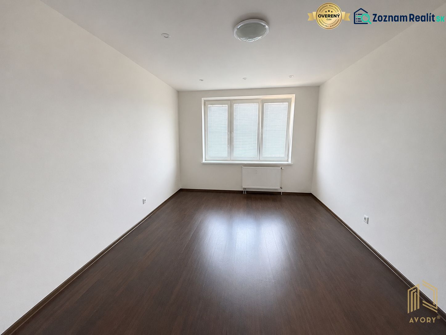 A room in a 2-room apartment with a wooden decor floor and three windows.
