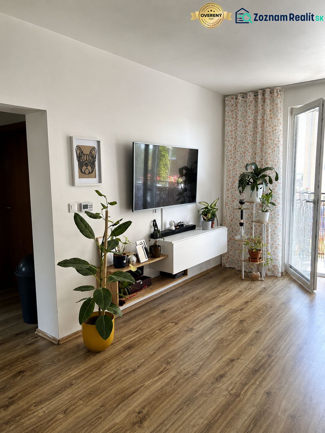 A room in a 3-room apartment with a wooden decor floor, a television, and plants.