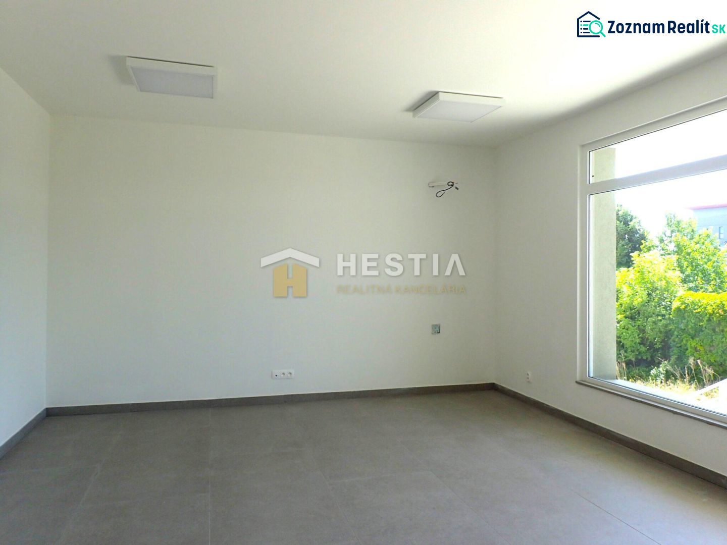 A spacious room with a large window, light-colored walls, and a floor with a wooden decor.