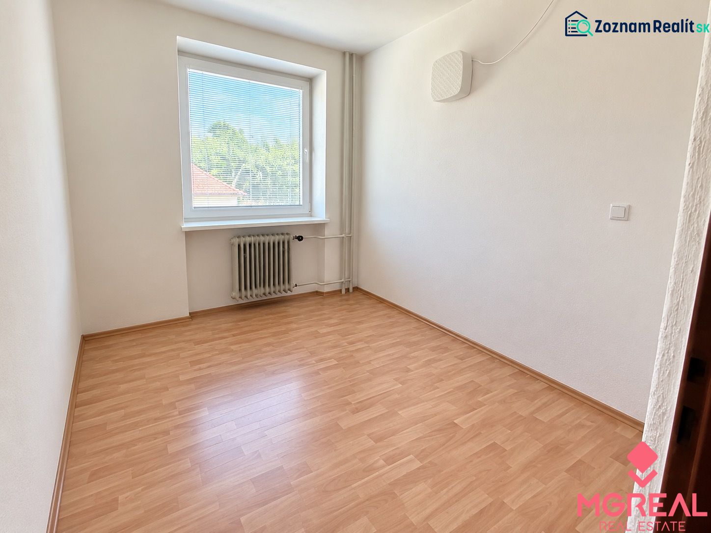 An empty room with a window, wooden floor, white walls, and a radiator.
