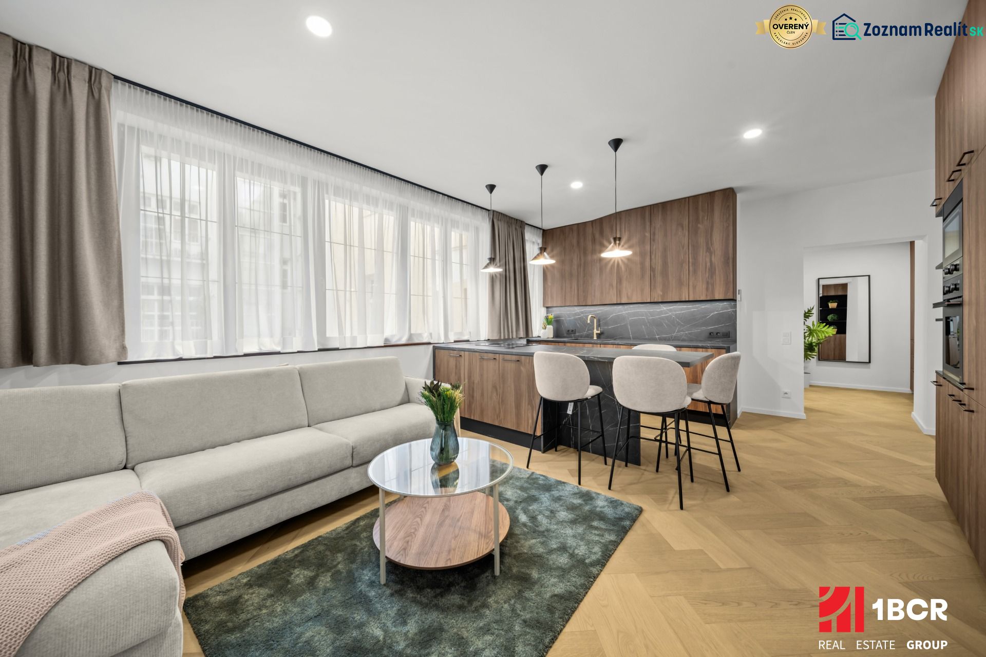 Living room in a two-room apartment with a sofa, kitchenette, and wooden decor flooring.