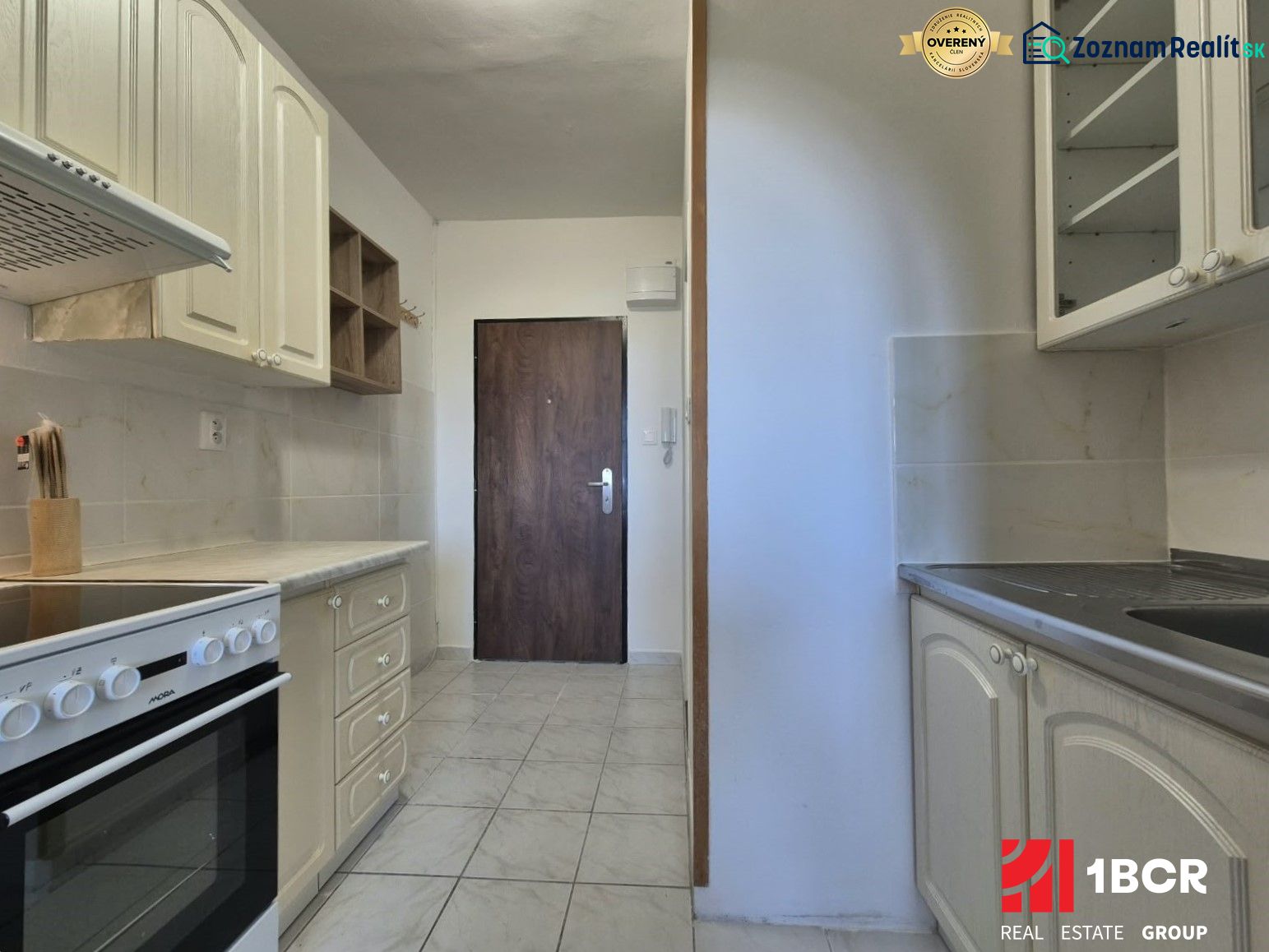 A kitchenette in a studio apartment with a white kitchen unit and ceramic flooring.