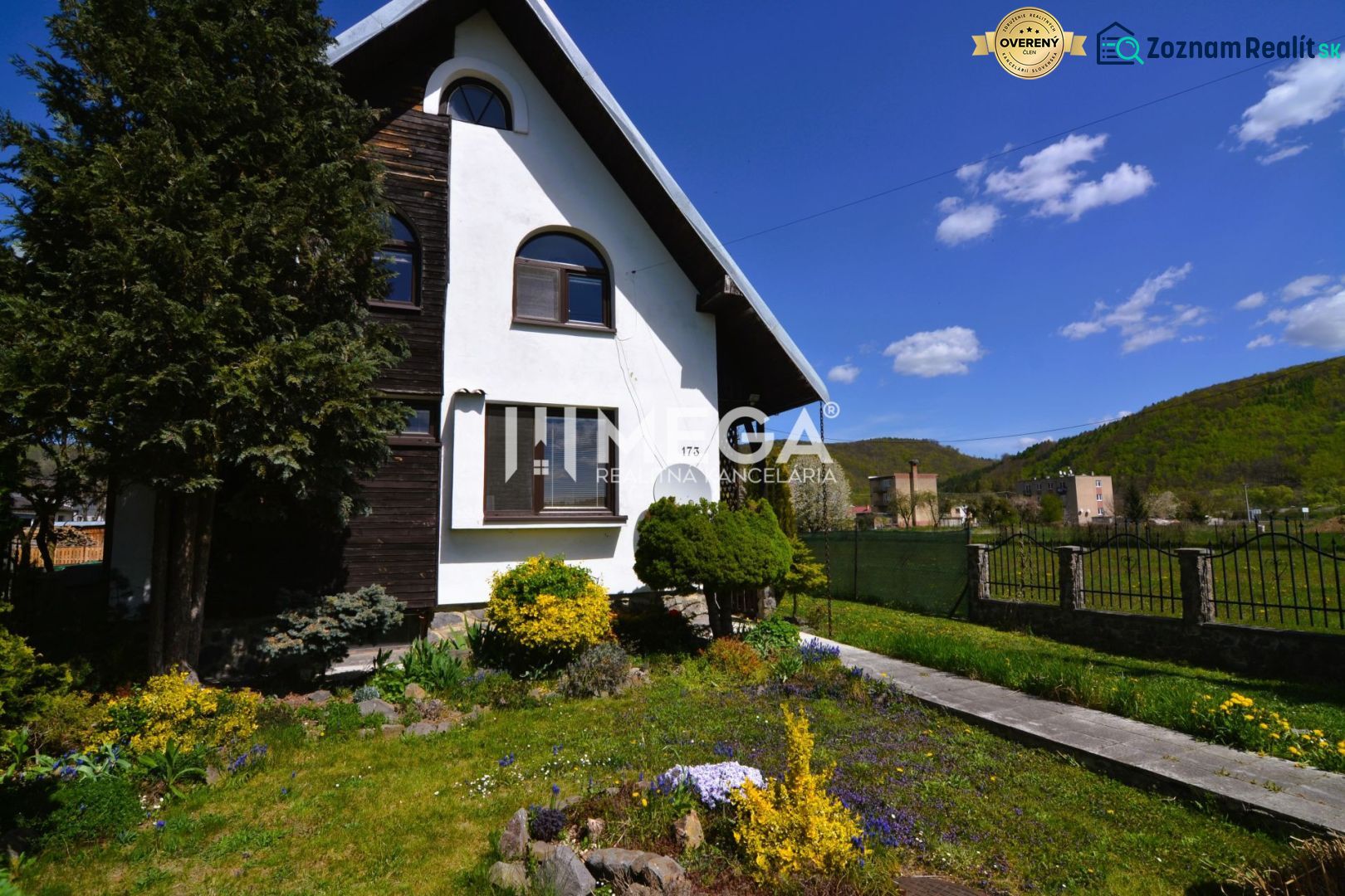 A family house in Koškovce with a landscaped garden and a hilly background under a blue sky.