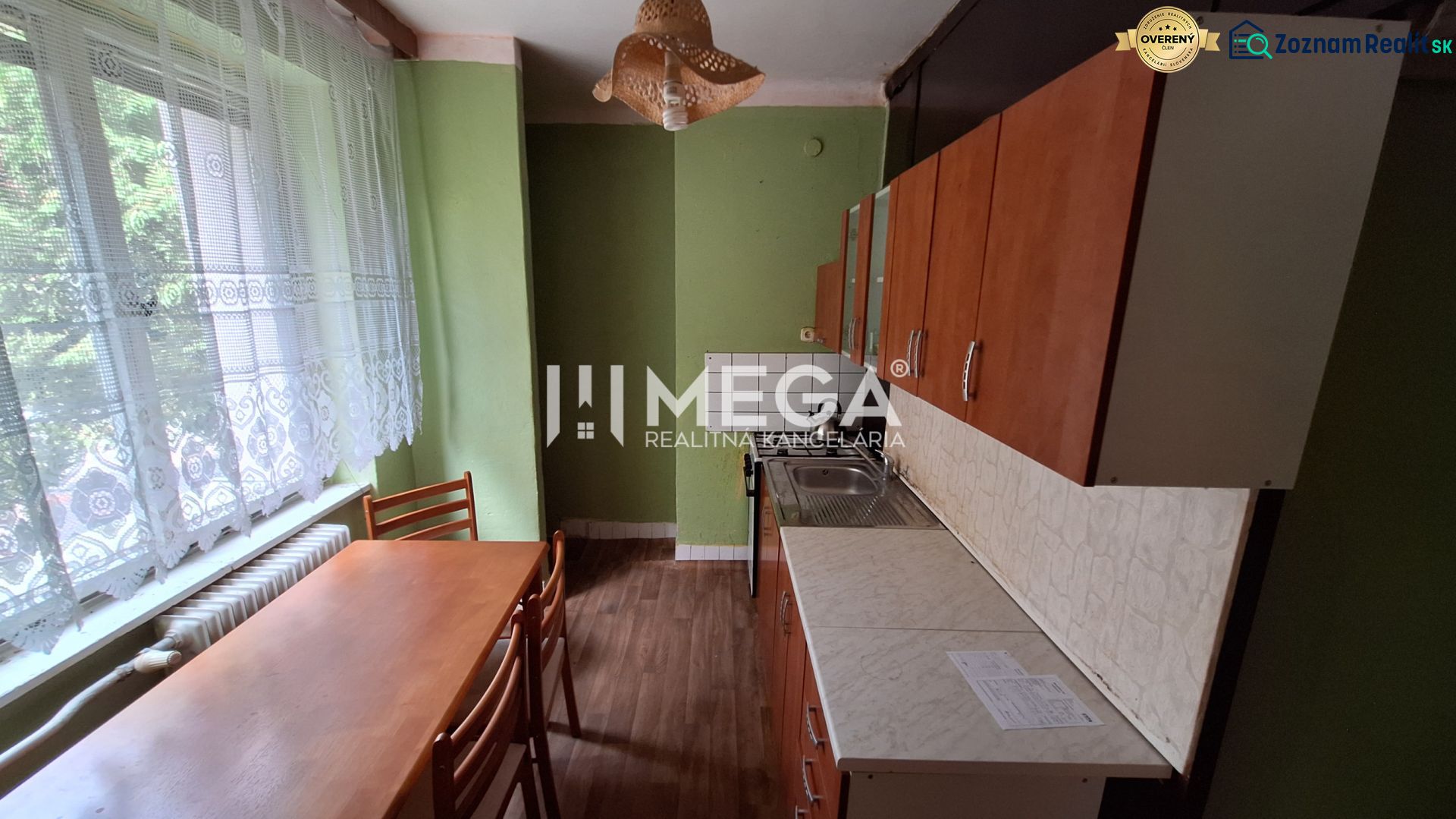 A kitchen in a 2-room apartment with a kitchen unit, table, wooden decor flooring, and curtains.