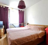 A bedroom in a three-room apartment with a bed, a television, and purple curtains.