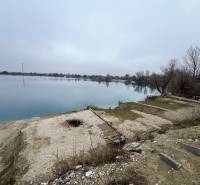 A view of a clear lake and natural surroundings at Veľké Košariská street. In the foreground, there are recreational plots with pebbles and a cold fire pit, while the surrounding landscape offers peace and quiet scenes typical of recreational areas.