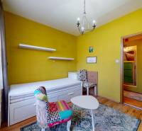 A room in a 3-room apartment with a bed, a chair, a table, and shelves on the wall.