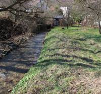 A stream with a grassy bank on Záhrady Street, surrounded by trees near a house, creates a peaceful view.