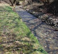 A stream surrounded by grass and trees, located on Myslavská and Záhrady streets.