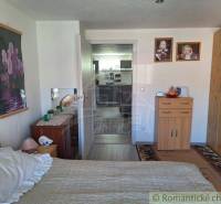 A bedroom in a family house with furniture and decorations, visible hallway and part of the kitchen.