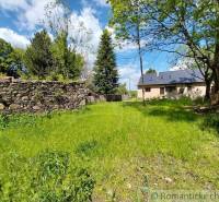 A cottage in Ábelová with a stone fence and a green lawn, surrounded by trees.