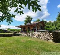 A cottage in Veľký Krtíš surrounded by nature, a stone wall, and a blue sky.