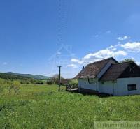 A cottage in Podkriváni surrounded by a green meadow and forests under a clear blue sky.
