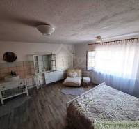 A bright room in a family house with a bed, a sink, a chair, and a large window.