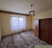 A room in a family house with a window curtain, old furniture, and an unfinished floor.