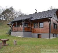 A cottage with wooden cladding, a gable roof, and a yard, located in hilly countryside.