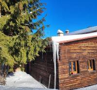 A wooden cottage with a glacier on the roof, a snowy landscape, and a family house in the background.