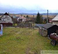 A rural garden with an old building, stacks of wood, hills in the distance.