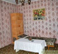 Interior of a family house: tiled stove, patterned wallpaper, sofa, table with tablecloth, painting of flowers.
