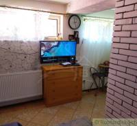 Television in the corner of the room next to the clock, masonry, furniture, blinds, and a family house.