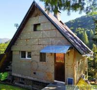 A cottage in a natural setting, sunlight illuminates the roof, surrounded by a garden and hills.