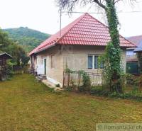 A family house with a red roof, greenery, and a hilly scenery in the background.