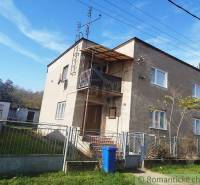 A two-story family house with a balcony, fencing, and a blue waste bin on a grassy plot.