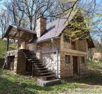 A stone cottage in the forest with an outdoor staircase and seating area, surrounded by trees in spring.
