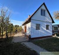 A simple white cottage with a bicycle, wooden door, trees, and a sunny background.