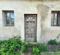 A country cottage with old wooden doors and two windows, surrounded by greenery.