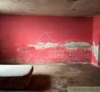 A shabby room with a red wall, a worn-out mattress; visible signs of dampness.