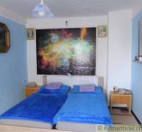 A bedroom with blue sheets, a colorful painting, nightstands, and pictures on the wall.