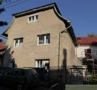 A two-story family house with a metal roof, an external staircase, and a parked green car.