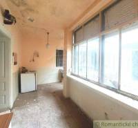 A veranda in an old cottage with large windows, a damaged ceiling, and a refrigerator.