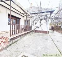 Family house with a terrace, fencing, and trees on a concrete yard.