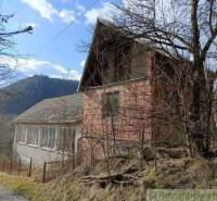 An old cottage in nature, surrounded by trees and a nearby mountainous environment.
