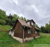 A wooden cottage in Veľké Rovné, surrounded by greenery and forests, under a cloudy sky.