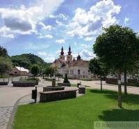 Square with a church and garden, trees, benches, hills and houses in the background.