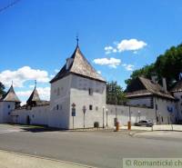 A medieval structure of towers and walls, with gardens in the background under a blue sky.